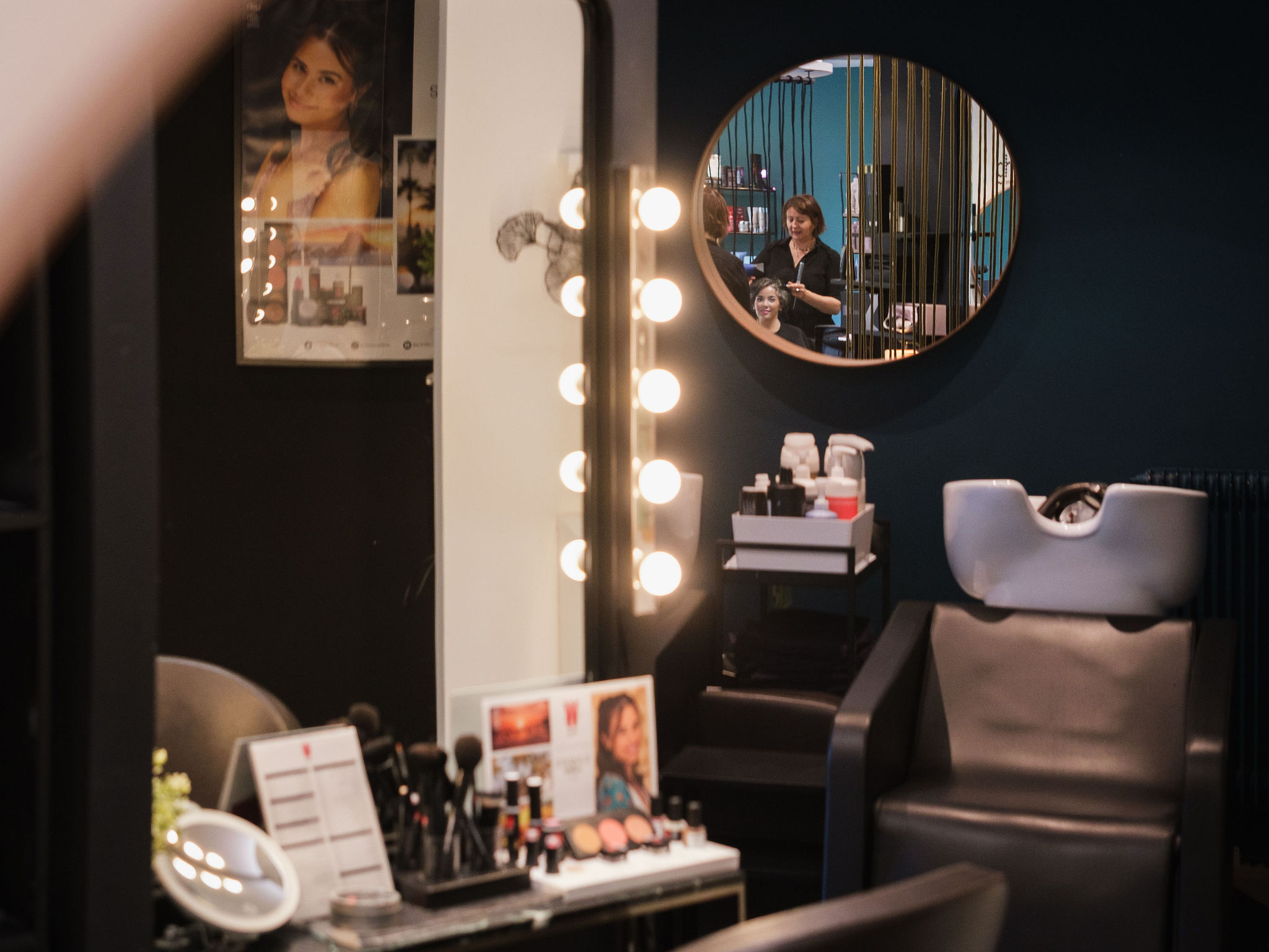 Hair salon interior with makeup station and a stylist working on a client's hair reflected in a round mirror.